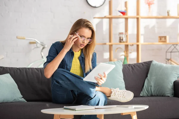 Konzentrierte blonde Frau mit Brille blickt auf digitales Tablet, während sie auf der Couch auf verschwommenem Hintergrund sitzt — Stockfoto