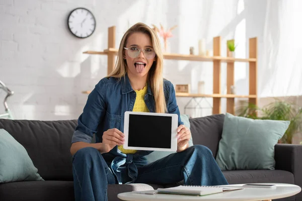 Mujer rubia joven con la lengua que sobresale mirando a la cámara mientras muestra la tableta digital en casa sobre un fondo borroso - foto de stock