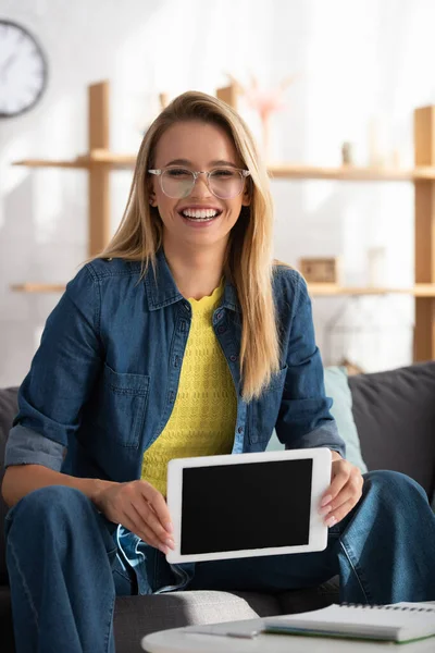 Fröhliche junge blonde Frau blickt in die Kamera, während sie zu Hause auf verschwommenem Hintergrund ein digitales Tablet zeigt — Stockfoto