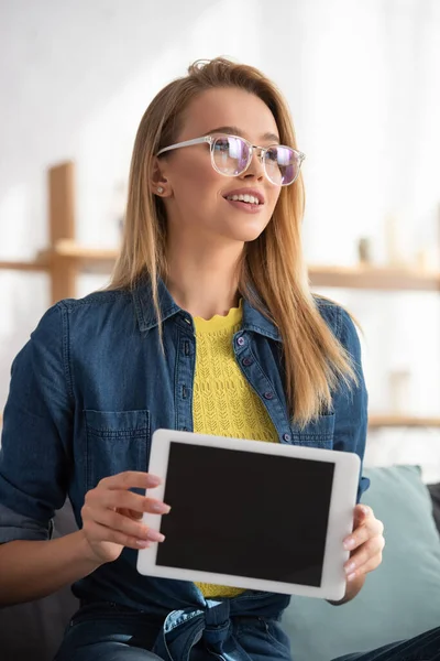 Positive blonde woman in eyeglasses looking away while showing digital tablet on blurred background — Stock Photo