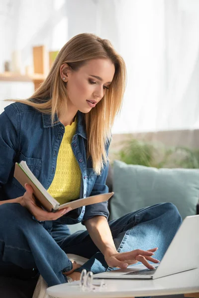 Young blonde woman with notebook typing on laptop at home on blurred background — Stock Photo