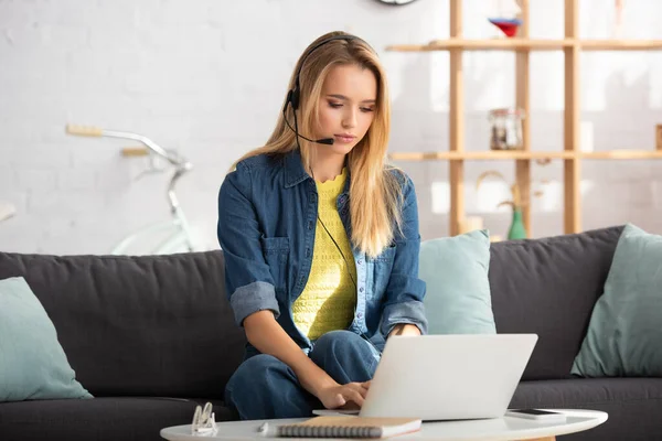Young blonde woman in headset typing on laptop while sitting on couch on blurred background — Stock Photo