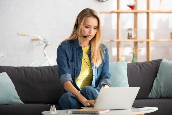 Mujer rubia joven en auriculares escribiendo en el ordenador portátil mientras está sentado en el sofá sobre un fondo borroso - foto de stock