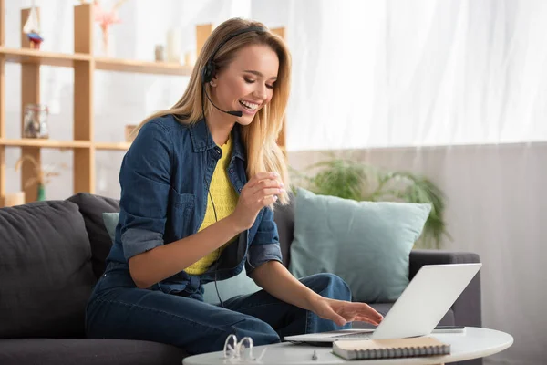 Allegro donna bionda in auricolare ridere mentre seduto sul divano vicino al computer portatile a casa su sfondo sfocato — Foto stock