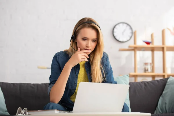 Nachdenkliche junge Frau mit Headset sitzt neben Laptop zu Hause auf verschwommenem Hintergrund — Stockfoto