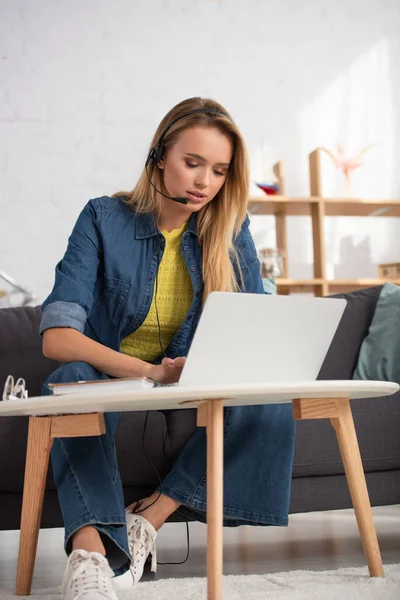 Молодая блондинка в наушниках печатает на ноутбуке, сидя на диване возле журнального столика на размытом фоне — стоковое фото