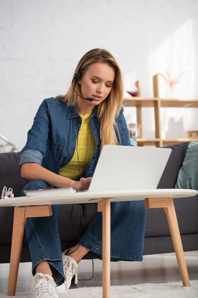 Jovem loira no fone de ouvido digitando no laptop enquanto estava sentado no sofá perto da mesa de café no fundo embaçado — Fotografia de Stock