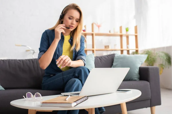 Young woman in headset sitting near coffee table with devices, notebook and eyeglasses at home on blurred background — Stock Photo