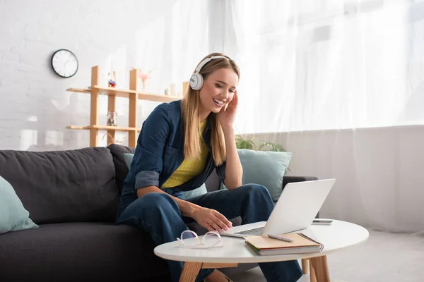 Cheerful young blonde woman in headphones looking at laptop while sitting on couch at home — Stock Photo