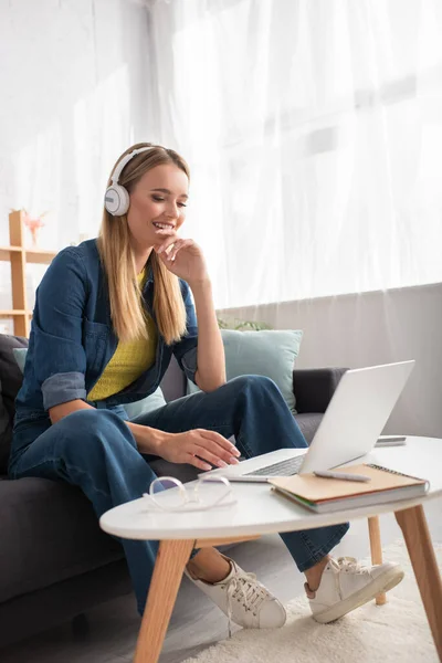 Glückliche junge Frau mit Kopfhörern, die auf Laptop schaut, während sie auf der Couch neben dem Couchtisch auf verschwommenem Hintergrund sitzt — Stockfoto