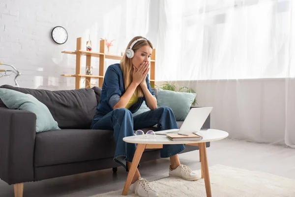 Full length of scared young woman in headphones looking at laptop while sitting on couch near coffee table — Stock Photo