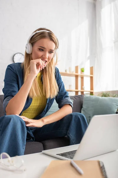 Занепокоєна молода жінка в навушниках дивиться на ноутбук, сидячи на дивані на розмитому передньому плані — стокове фото