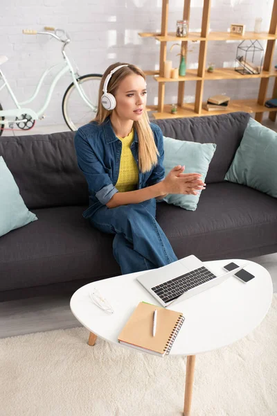 Hochwinkelaufnahme einer blonden Frau mit Kopfhörern, die wegschaut, während sie auf einer Couch in der Nähe eines Couchtisches mit digitalen Geräten sitzt — Stockfoto