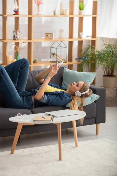 Junge blonde Frau mit Kopfhörer textet auf dem Smartphone, während sie zu Hause auf dem Sofa neben dem Couchtisch liegt — Stockfoto