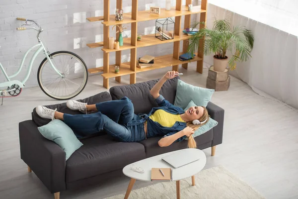 Blick aus dem Hochwinkel auf eine glückliche blonde Frau mit Kopfhörern, die lacht, während sie zu Hause auf der Couch liegt — Stockfoto