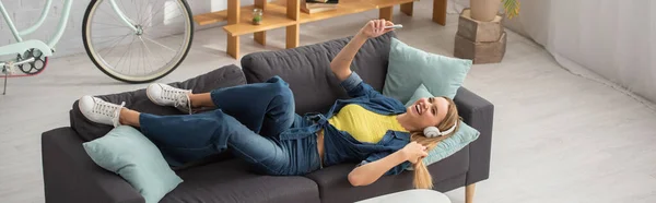 Vista de ángulo alto de la mujer rubia feliz con teléfono inteligente riendo mientras está acostado en el sofá en casa, pancarta - foto de stock