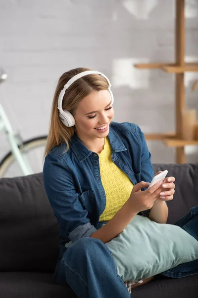 Smiling blonde woman in headphones texting on smartphone at home on blurred background — Stock Photo