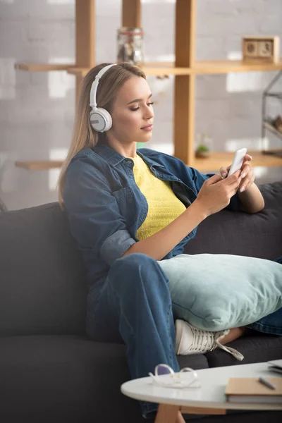 Junge blonde Frau mit Kopfhörer, die auf dem Smartphone SMS schreibt, während sie auf der Couch vor verschwommenem Hintergrund sitzt — Stockfoto