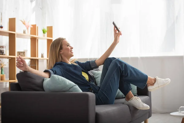 Ganzer Länge lächelnde blonde Frau mit Smartphone, die ein Selfie macht, während sie auf der Couch auf verschwommenem Hintergrund sitzt — Stockfoto