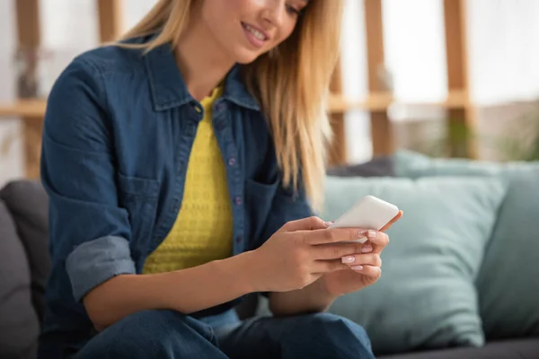 Vue recadrée de la jeune femme blonde texter sur smartphone tout en étant assis sur le canapé sur fond flou — Photo de stock