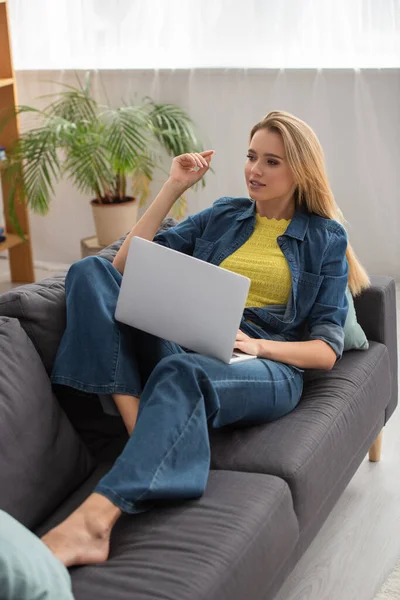 Полная длина молодой женщины, отводящей взгляд, пользуясь ноутбуком на диване дома — стоковое фото