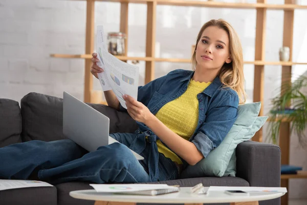 Junge blonde Frau mit Papieren blickt in die Kamera, während sie auf der Couch in der Nähe digitaler Geräte auf verschwommenem Hintergrund sitzt — Stockfoto