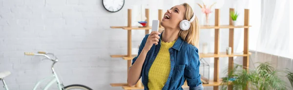 Mujer joven feliz en auriculares cantando mientras sostiene el teléfono inteligente en casa sobre fondo borroso, pancarta - foto de stock