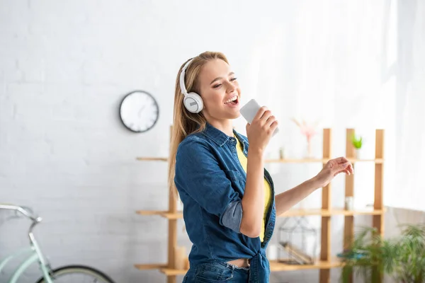 Fröhliche blonde Frau mit Kopfhörern singt, während sie ihr Smartphone zu Hause auf verschwommenem Hintergrund hält — Stockfoto