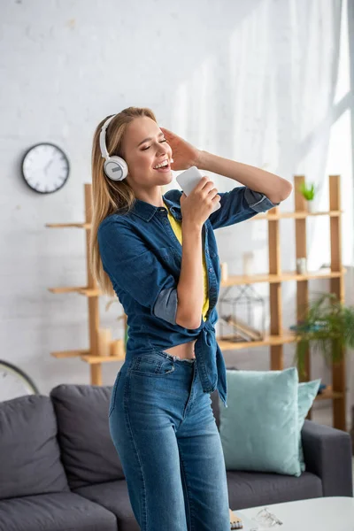 Happy blonde woman in headphones singing while holding smartphone at home on blurred background — Stock Photo