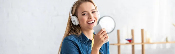 Усміхнена молода жінка в навушниках співає, тримаючи смартфон вдома на розмитому фоні, банер — стокове фото