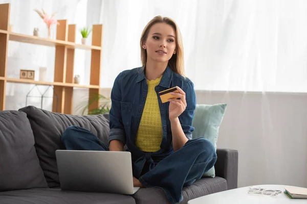 Молода блондинка з кредитною карткою дивиться далеко, сидячи біля ноутбука на дивані на розмитому фоні — стокове фото