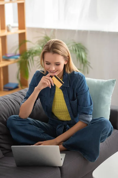Lächelnde junge blonde Frau mit Kreditkarte schaut auf Laptop, während sie auf Couch auf verschwommenem Hintergrund sitzt — Stockfoto