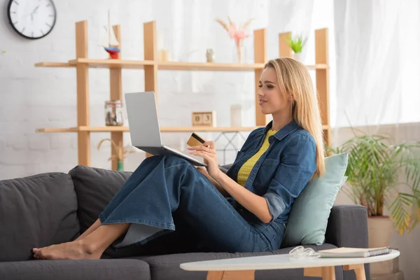 Ganzer Länge lächelnde blonde Frau mit Kreditkarte, die auf Laptop schaut, während sie auf Couch auf verschwommenem Hintergrund sitzt — Stockfoto