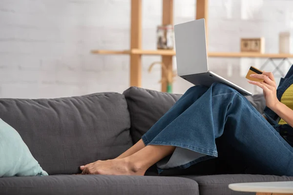 Vista parcial de la mujer joven con el ordenador portátil y la tarjeta de crédito sentado en el sofá en casa sobre fondo borroso — Stock Photo