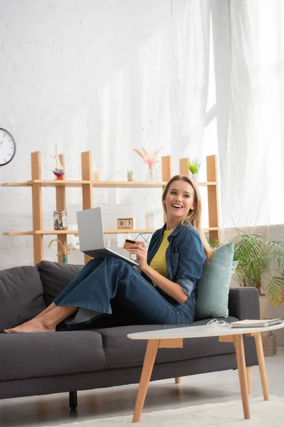 Happy blonde woman with credit card and laptop looking away while sitting on couch near coffee table on blurred background — Stock Photo