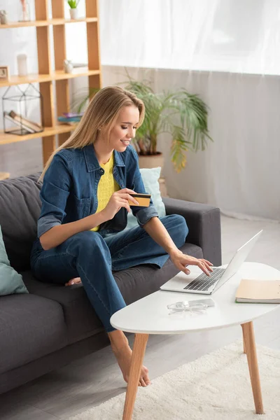 Full length of happy blonde woman with credit card typing on laptop at home on blurred background — Stock Photo