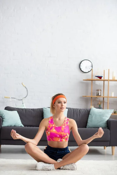 Full length of young sportswoman sitting in lotus pose and looking away at home on blurred background — Stock Photo