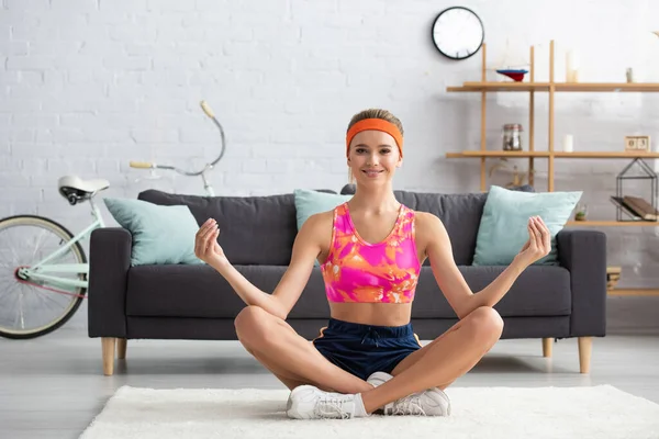 Full length of smiling sportswoman looking at camera while sitting in lotus pose at home on blurred background — Stock Photo