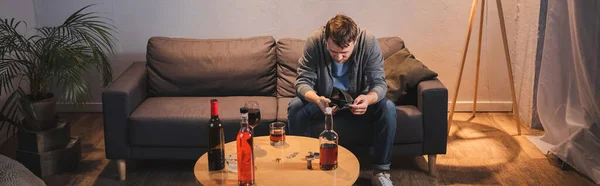 Hombre borracho sosteniendo cartera vacía cerca de la mesa con botellas de bebidas alcohólicas en casa, pancarta - foto de stock