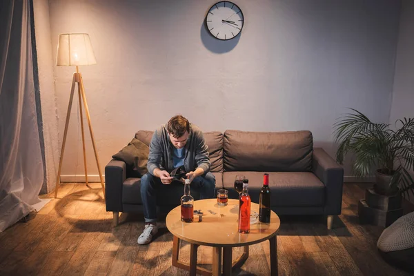 Alkoholsüchtiger Mann hält leere Brieftasche mit Flaschen am Tisch — Stockfoto