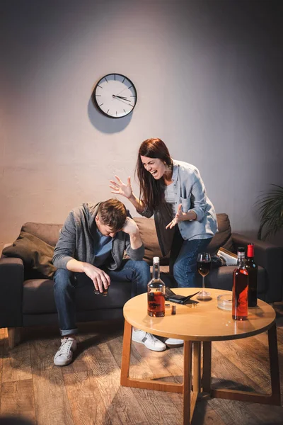 Mujer enojada gritando al marido borracho cerca de la mesa con bebidas alcohólicas en casa - foto de stock