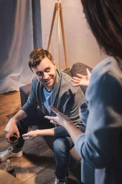 Drunk, smiling man holding glass of whiskey while looking at wife quarreling on blurred foreground — Stock Photo