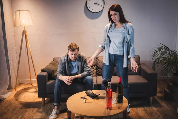 Verärgerte Frau steht mit offenen Armen neben betrunkenem Ehemann und Tisch mit Flaschen und leerem Portemonnaie — Stockfoto