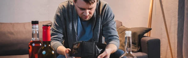 Hombre deprimido, borracho sosteniendo la cartera vacía cerca de botellas de alcohol en primer plano borrosa, pancarta - foto de stock