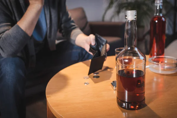 Vista recortada del hombre adicto sosteniendo la cartera vacía cerca de botellas con alcohol, fondo borroso - foto de stock