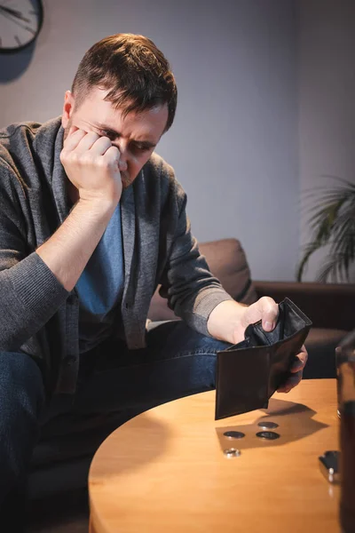 Hombre adicto al alcohol sosteniendo la cartera vacía cerca de monedas en la mesa - foto de stock