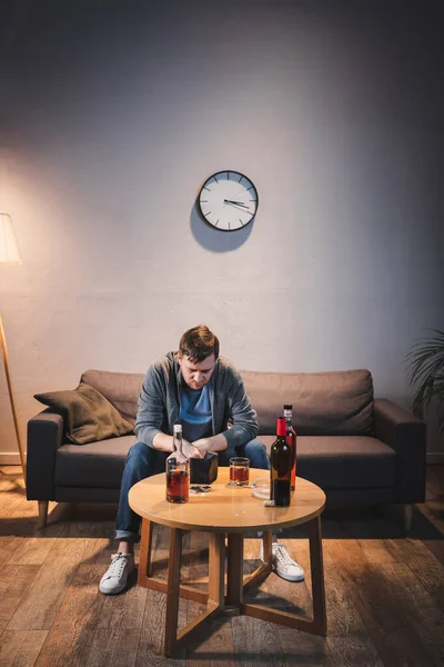 Depressed man holding empty wallet while sitting near bottles of alcohol at home — Stock Photo
