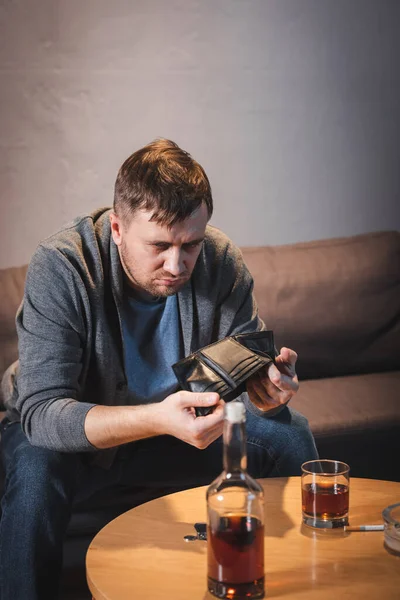 Uomo ubriaco con portafoglio vuoto vicino a bottiglia e bicchiere di whisky a casa — Foto stock