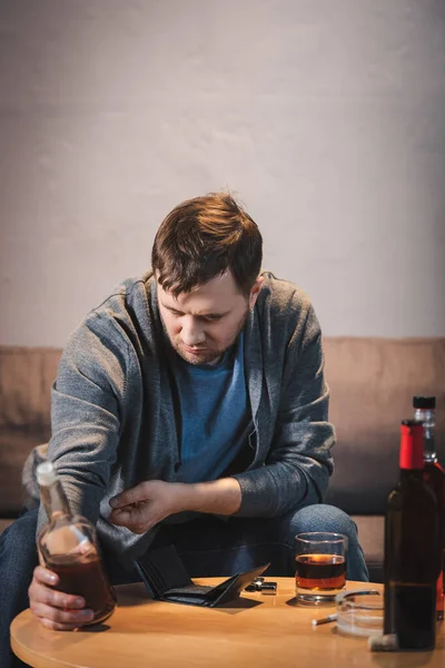 Frustrated, addicted man holding bottle of whiskey near empty wallet on table — Stock Photo