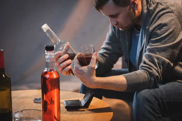 Hombre borracho sosteniendo botella y vaso de whisky cerca de la billetera vacía en la mesa - foto de stock